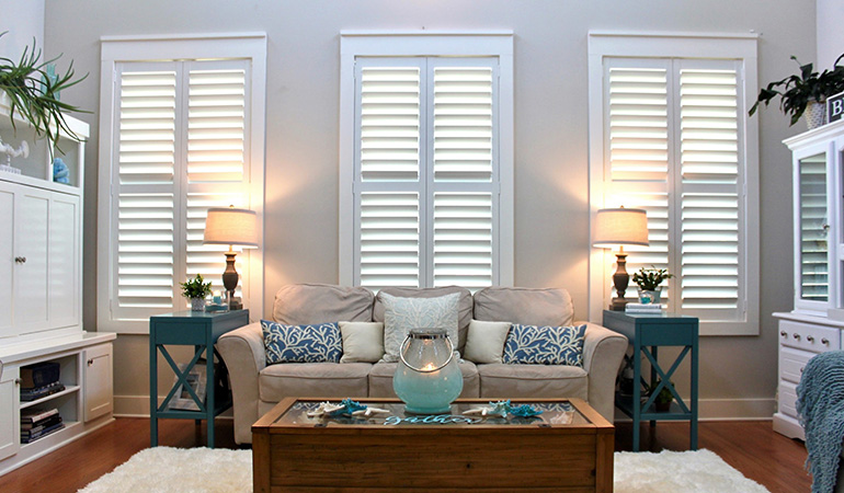 Energy-efficient Polywood shutters can reduce your energy bills. White Polywood shutters on three windows in a white and blue living room